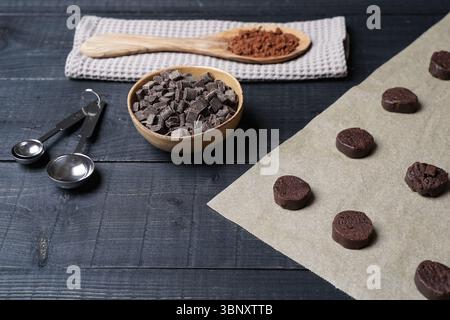 Vista frontale dell'impasto tagliato su carta da forno con cucchiaio di cacao sull'asciugamano da cucina, ciotola con gocce di cioccolato e misurini in metallo Foto Stock