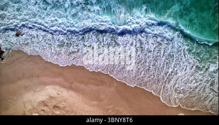 Le onde turchesi dell'oceano si schiantano su una sabbia liscia e abbronzante, formando una schiuma bianca di lacy dal design piatto Foto Stock