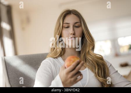 Giovane donna bionda con un top bianco si siede rilassata su una poltrona con una mela Foto Stock
