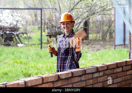 Una giovane muratrice con un casco arancione con un martello da costruzione e un mattone in mano sorride accanto a un muro di mattoni Foto Stock