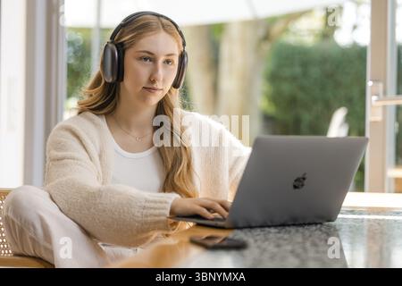 Giovane donna bionda con la parte superiore bianca si siede rilassata al tavolo con le cuffie e il laptop Foto Stock