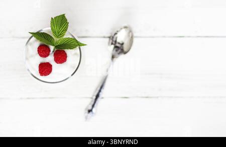 Un bicchiere con yogurt al latte, lamponi maturi e una foglia verde su un tavolo di legno bianco con una vista dall'alto dello spazio copia, fuoco selettivo Foto Stock