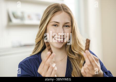 Giovane donna bionda con 2 pezzi di cioccolato, sorridente Foto Stock