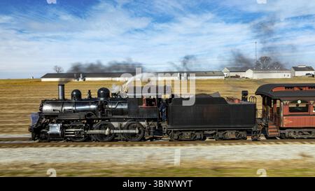 Una locomotiva a vapore d'epoca alimenta lungo i binari, inviando pennacchi di fumo nel cielo blu. I campi circostanti e le case coloniche ne esaltano il pittoresco Foto Stock