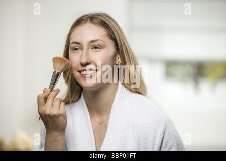 Giovane donna bionda in accappatoio bianco sorridente, con spugnetta in polvere, sorridente Foto Stock
