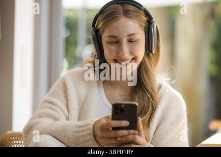 Giovane donna bionda con la parte superiore bianca si siede rilassata al tavolo con le cuffie e il telefono cellulare, sorridendo Foto Stock