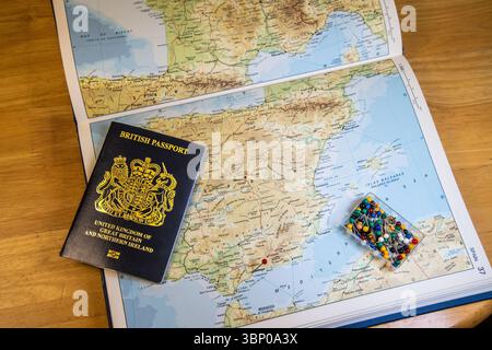 Passaporto britannico di nuovo stile su un atlante aperto alla Spagna, con una spilla rossa che indica Málaga e un pacchetto di spille colorate nelle vicinanze, che simboleggiano la pianificazione del viaggio Foto Stock