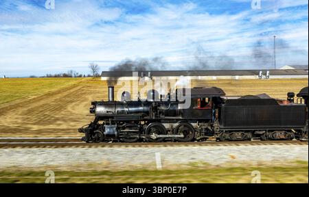 Una locomotiva a vapore d'epoca alimenta lungo i binari, inviando pennacchi di fumo nel cielo blu. I campi circostanti e le case coloniche ne esaltano il pittoresco Foto Stock