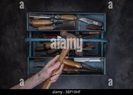 Vista dall'alto verso il basso di una mano che regge un vecchio martello con manico in legno sopra una cassetta degli attrezzi aperta in metallo blu riempita di utensili su una superficie scura e ruvida Foto Stock