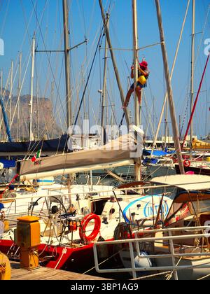 Uomo che arrampica sull'albero di barche a vela, porto turistico di Palermo pieno di yacht attraccati, scenario del porto estivo della Sicilia Foto Stock