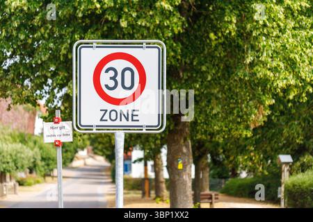 Un segnale tedesco del limite di velocità "30 zone" è ben visibile su uno sfondo di alberi verdi che costeggiano una strada in un'area residenziale.n Foto Stock