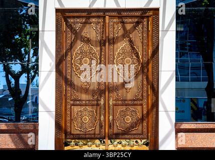 Una porta di legno splendidamente realizzata con intricati intagli e motivi floreali, adagiata su una facciata moderna di un edificio con riflessi in vetro. Foto Stock