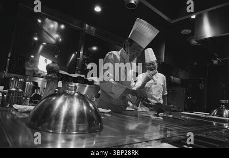 Due cuochi sorridenti teppanyaki preparano il cibo alla griglia, creando un'esperienza culinaria calda e invitante Foto Stock