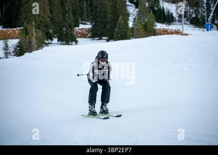 Un uomo sta sciando su una pista innevata con una macchina fotografica in mano. Indossa un vestito bianco e nero e un casco Foto Stock