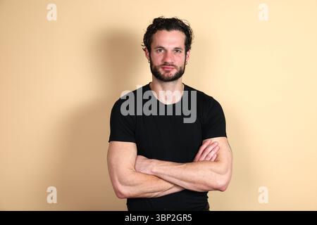 Un giovane con i capelli scuri e la barba, che indossa una t-shirt nera, sta con le braccia incrociate, guardando con sicurezza e direttamente alla telecamera. Studio Foto Stock