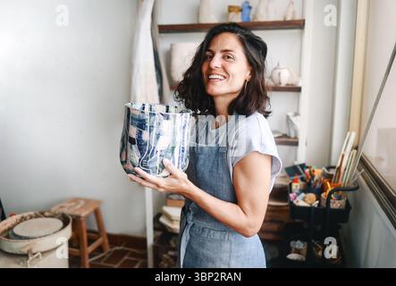 Donna latina artista di ceramica che tiene un vaso dipinto in studio di ceramica, mostrando lavori fatti a mano Foto Stock