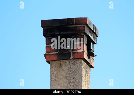 Un camino in mattoni con una parte superiore annerita e macchiata di fuliggine si innalza contro un cielo blu limpido, mostrando segni di uso e esposizione a lungo termine Foto Stock