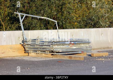 Una pila ordinata di tubi per impalcature in metallo galvanizzato poggia su tavole di legno accanto a una barriera in calcestruzzo in un cantiere stradale, con alberi Foto Stock