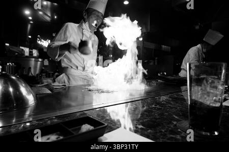 Lo chef accende fiamme spettacolari su una griglia teppanyaki, attraenti commensali in una scena in bianco e nero Foto Stock