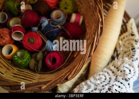 I cestelli in tessuto contengono bobine e palline di filo vivaci in vari colori, tra cui rosso, verde e rosa. Un cono di filettatura più grande giace accanto al tessuto Foto Stock