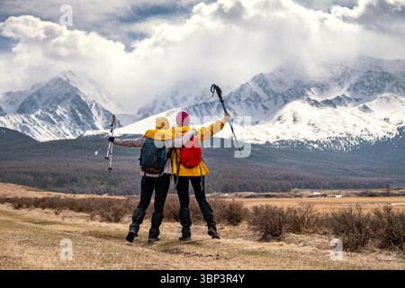 Due escursioniste con zaini si arroccano e sollevano le braccia ai lati con pali da trekking sullo sfondo di alte cime innevate dei Monti Altai, vista dal retro Foto Stock