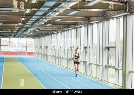 Atleta femminile che corre su pista blu in erba con scarpe da running sotto le travi della finestra, spazio copia Foto Stock