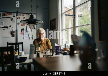 Donna anziana contemplativa seduta al tavolo da pranzo a casa Foto Stock