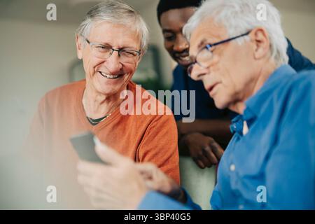 Uomo anziano che condivide lo smartphone con un amico e un infermiere a casa di riposo Foto Stock