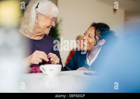 Donna sorridente esperta di assistenza sanitaria accovacciata vicino anziana donna maglieria a casa di riposo Foto Stock