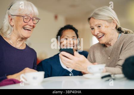Donna anziana sorridente che condivide lo smartphone con un amico e un infermiere a casa di riposo Foto Stock