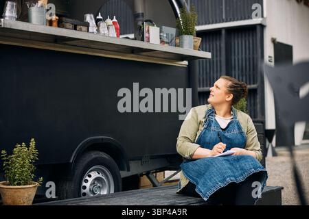 Giovane donna seduta su una tavola mentre scrive appunti vicino al furgone del cibo Foto Stock