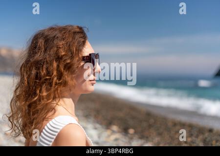 Donna occhiali da sole Beach: Giovane donna con occhiali da sole che guarda in mare sulla spiaggia rocciosa durante il giorno. Foto Stock