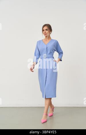Donna elegante in blu, vestito con cintura, cammina con sicurezza in avanti, una mano in tasca, l'altra leggermente sollevata. Indossa tacchi rosa brillanti, capelli legati Foto Stock