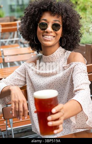 Giovane donna nera con occhiali da sole che tiene una pinta di birra e sorride in un pub all'aperto Foto Stock