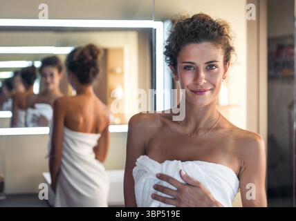 Una bella donna sorride e si avvolge in un asciugamano bianco dopo un bagno rilassante in un bagno moderno Foto Stock
