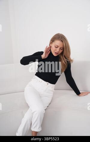 Una giovane donna con lunghi capelli biondi in un maglione nero e pantaloni bianchi si siede su un divano, guardando in basso, con la mano destra che le tocca il collo e la sinistra Foto Stock