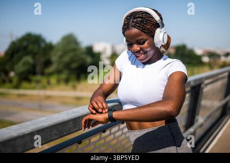 Donna africana felice in abbigliamento sportivo con cuffie che controllano il polso e il tempo con un orologio da polso intelligente dopo l'esercizio fisico sul ponte cittadino. Foto Stock