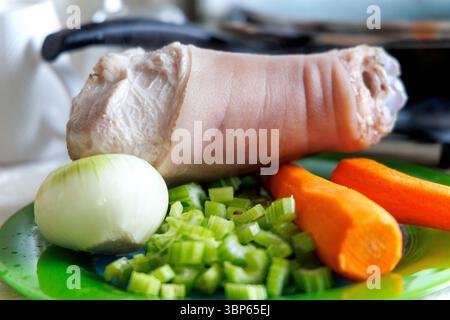 Uno stinco di maiale sbucciato è visibile su una superficie verde, circondato da verdure fresche: Steli di ​​celery affettati, mezza cipolla e due carote. Foto Stock
