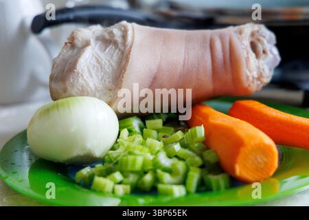 Uno stinco di maiale sbucciato è visibile su un piatto verde, accanto ad esso c'è mezza cipolla, gambi di sedano tritati e due carote. Foto Stock