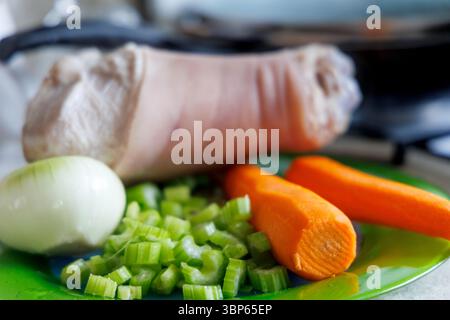 Uno stinco di maiale sbucciato è visibile su un piatto verde, accanto ad esso c'è mezza cipolla, gambi di sedano tritati e due carote. Foto Stock