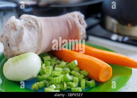 Uno stinco di maiale sbucciato è visibile su un piatto verde, accanto ad esso c'è mezza cipolla, gambi di sedano tritati e due carote. Foto Stock