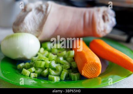 Uno stinco di maiale sbucciato è visibile su un piatto verde, accanto ad esso c'è mezza cipolla, gambi di sedano tritati e due carote. Foto Stock
