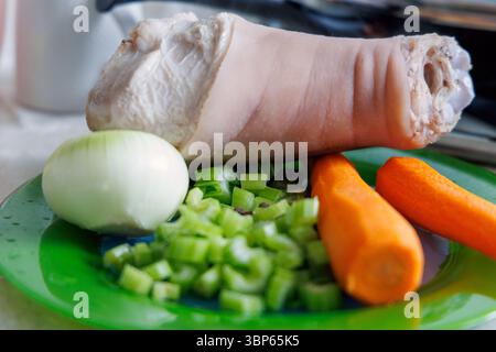 Uno stinco di maiale sbucciato è visibile su una superficie verde, circondato da verdure fresche: Steli di ​​celery affettati, mezza cipolla e due carote. Foto Stock