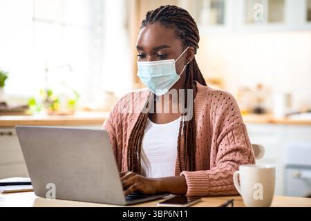 Giovane donna di affari nera che lavora da casa con un computer portatile, indossando una maschera medica Foto Stock