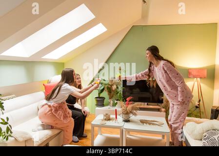 Amici vestiti con intimi indumenti da sonno che condividono le bevande e si godono un rilassante incontro al chiuso in un ambiente elegante e informale con illuminazione naturale a. Foto Stock