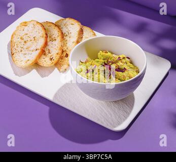 Cibo, ciotola di guacamole con fiori viola e fette di pane tostato su un piatto bianco su sfondo viola Foto Stock