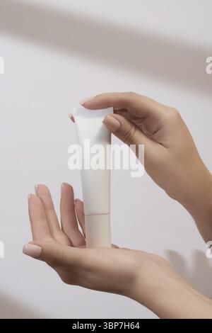 La mano della donna presenta un modello di un tubo di plastica bianco di crema per le mani. Confezione non etichettata per cosmetici alla luce del sole su sfondo bianco. Concetto di sk Foto Stock