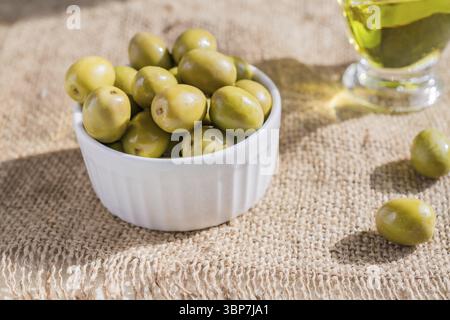 Olive verdi marinate fresche in una ciotola di ceramica bianca e una sauceboat di olio d'oliva vergine di prima qualità su fondo in tela di burlap Foto Stock
