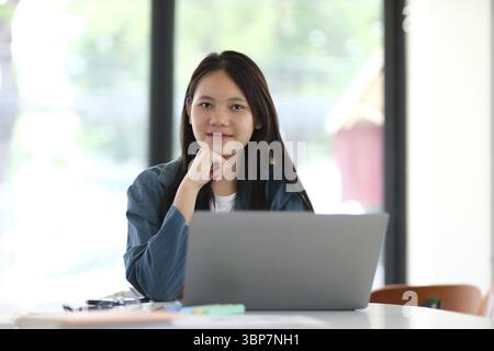 Una donna è seduta a un tavolo con un portatile di fronte a lei. Indossa una giacca blu e ha le mani in ginocchio Foto Stock