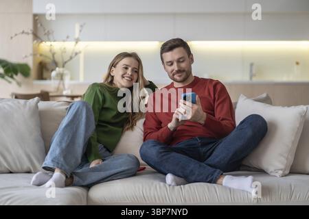 Coppia di sposi contenta che si rilassa sul divano di casa, l'uomo mostra lo schermo dello smartphone della moglie, pianifica un viaggio insieme, acquista online tramite telefono. Allegro marito Foto Stock